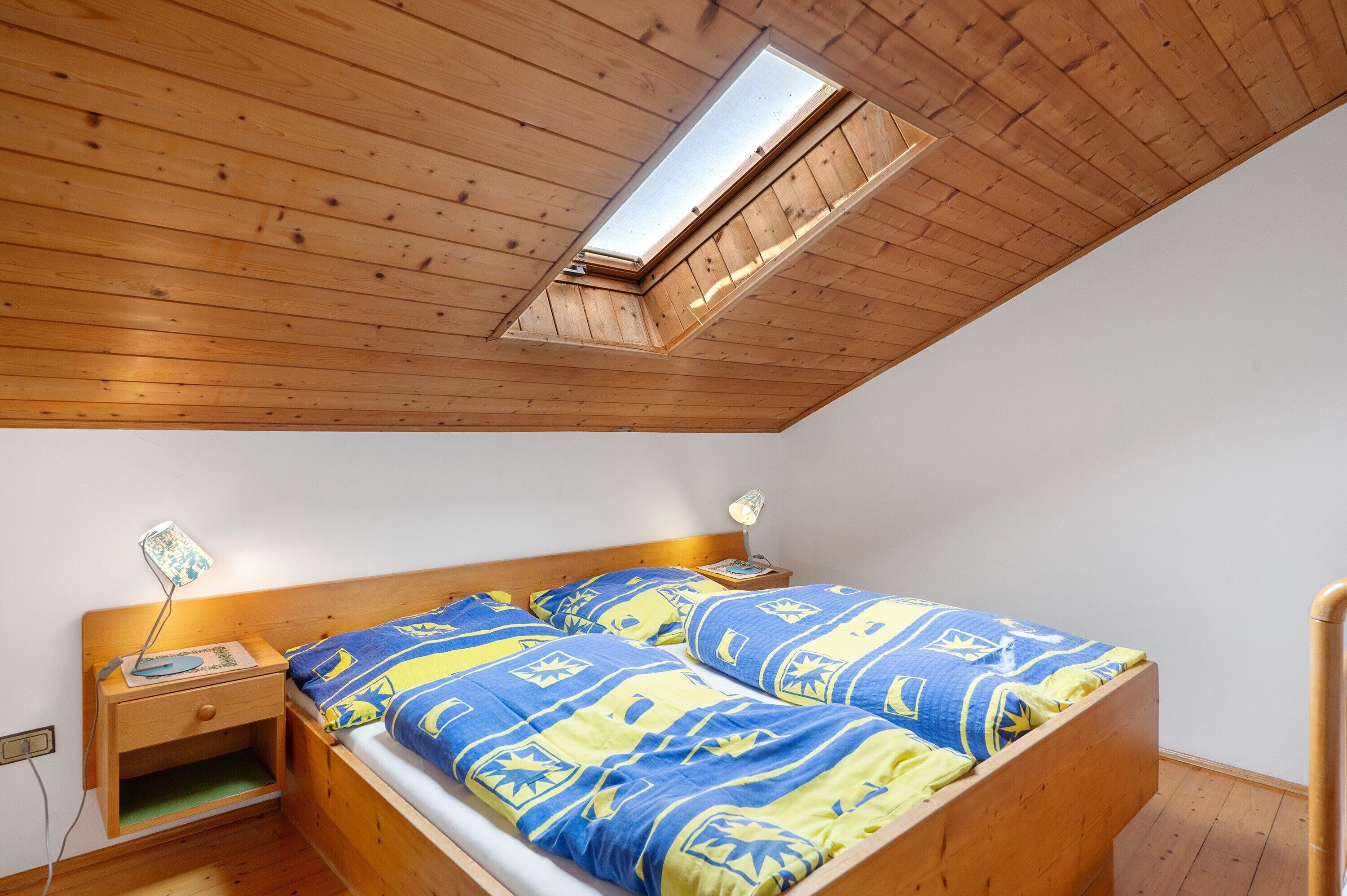 A bedroom with a sloped ceiling, a skylight, a wooden bed, a bedside table with a lamp, and a nightstand.