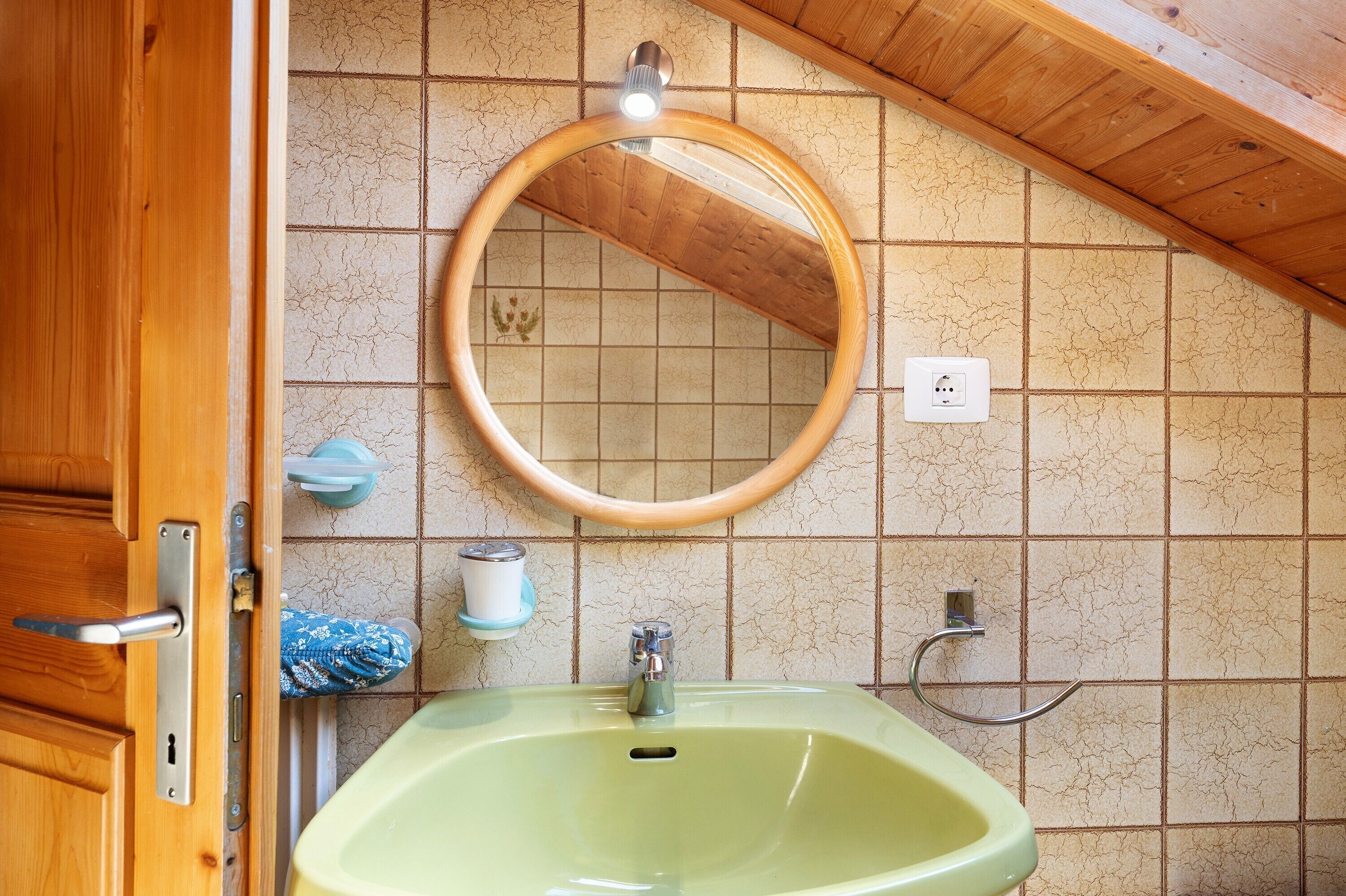 Un baño con puerta de madera, lavamanos verde, espejo redondo y un perchero para toallas.