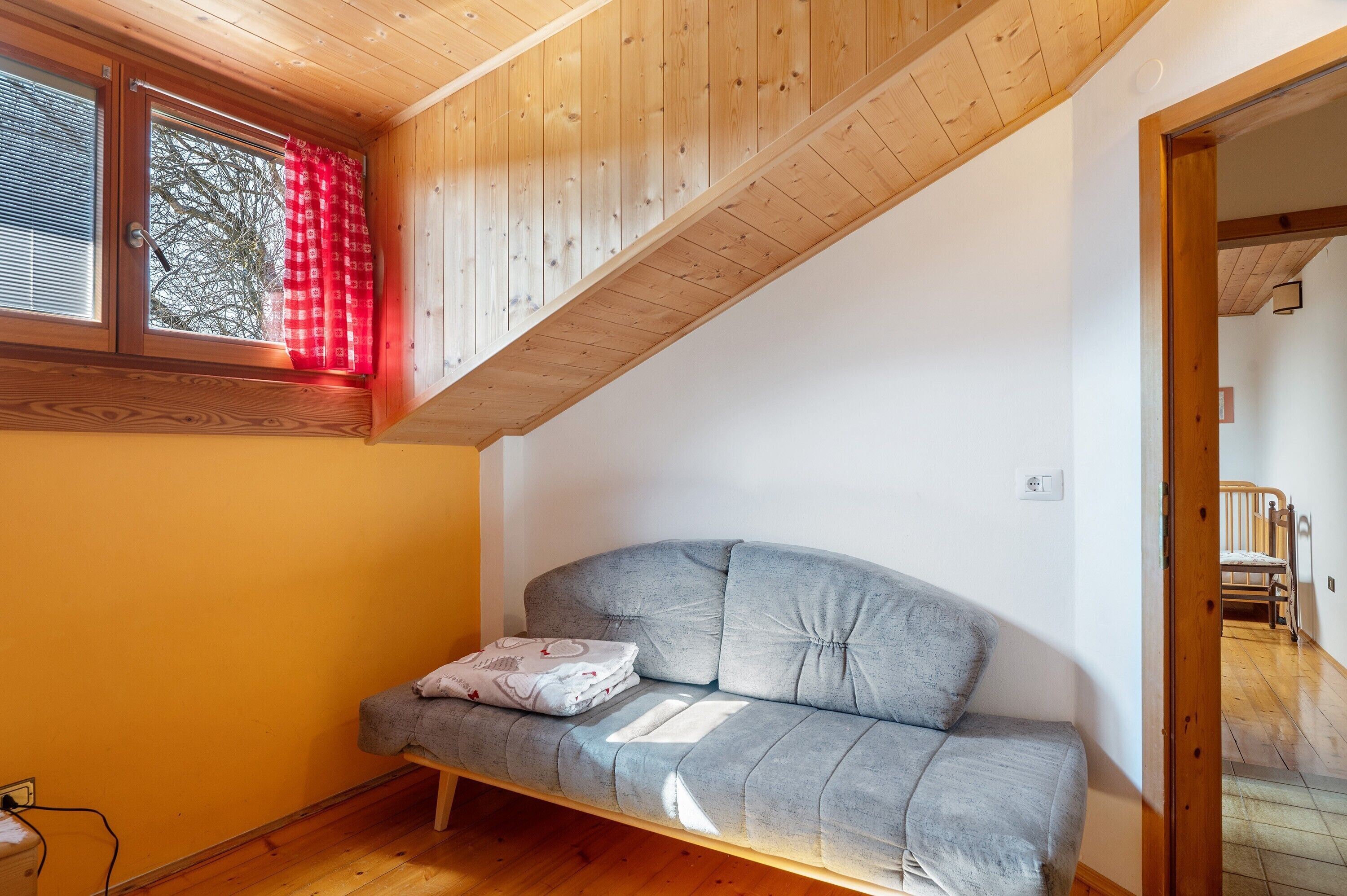 Ein Zimmer mit Holzbalkendecke, einem grauen Sofa mit Kissen, einem Fenster mit rotem Vorhang und Holzboden.