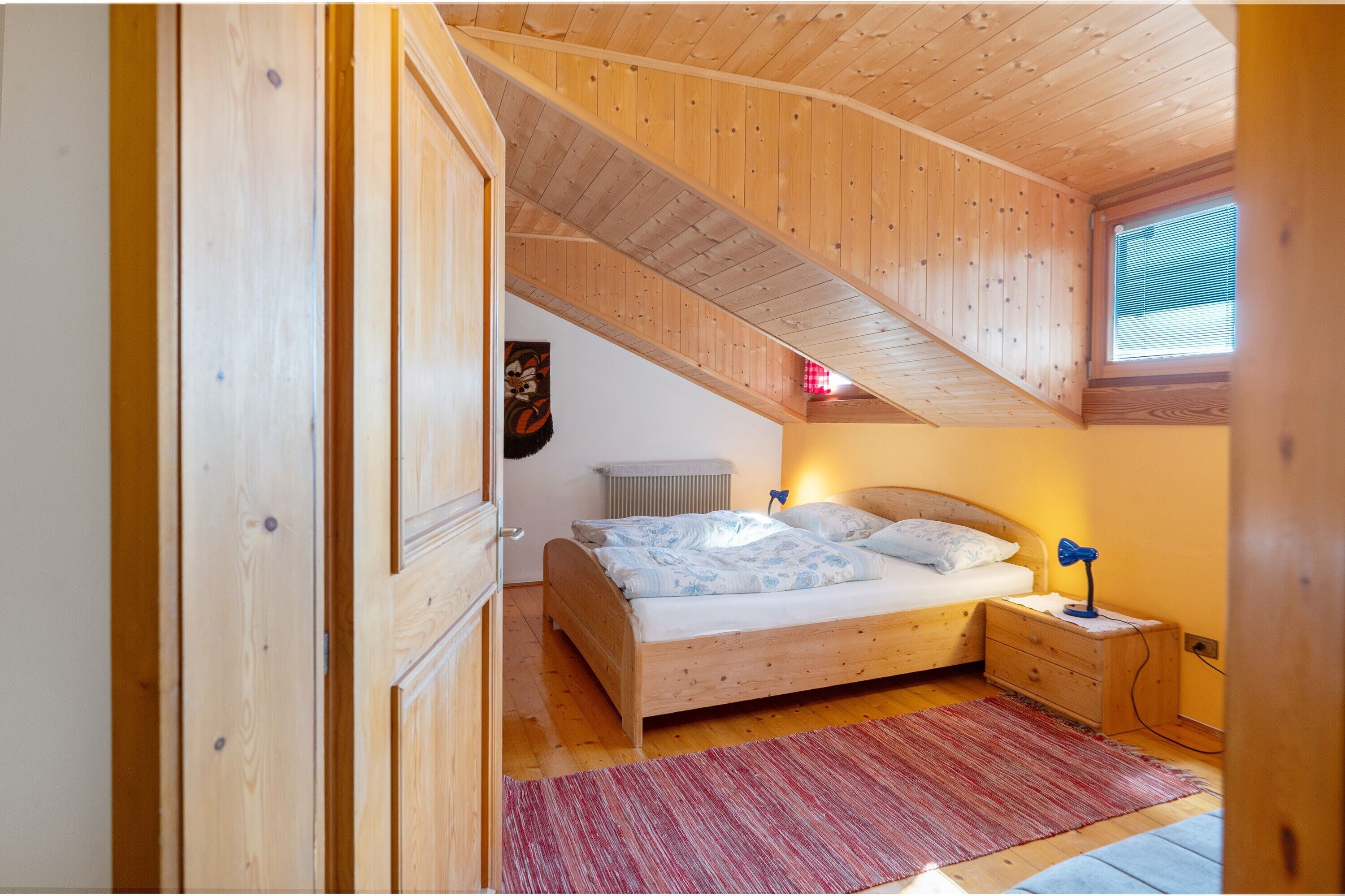 Une chambre avec un plafond en bois, un lit recouvert d’une couvre-lit à motifs blancs et bleus, un tapis rouge et une table de chevet avec une lampe.