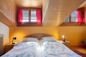 Une chambre avec un plafond en bois, deux fenêtres ornées de rideaux rouges, un lit avec une literie à motifs floraux, une lampe de chevet et une table de chevet.