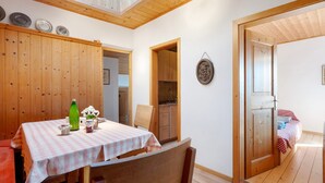 Un coin repas avec une table dressée pour deux personnes, des murs en bois, une cuisine visible à travers une porte ouverte et un lit dans la pièce adjacente.