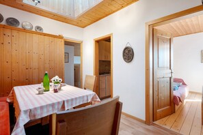 Comedor con una mesa lista para dos, paredes de madera, una cocina visible a través de una puerta abierta y una cama en la habitación contigua.