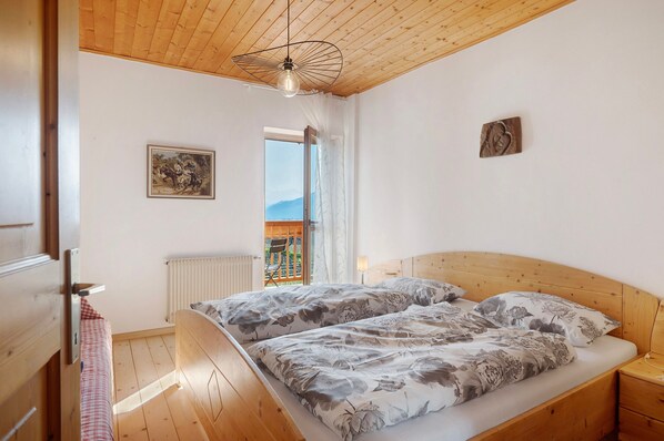 Un dormitorio con techo de madera, una cama con ropa de cama floral, una mesita de noche, un radiador y una ventana con vistas.