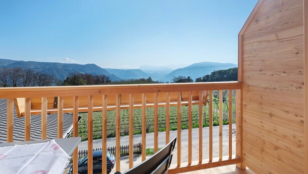 Un balcon avec une balustrade en bois, une table recouverte d’une nappe et une vue sur un vignoble et des montagnes.