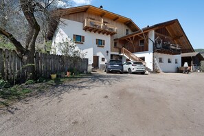 Un edificio de dos pisos con balcón de madera, camino de tierra y coches estacionados.