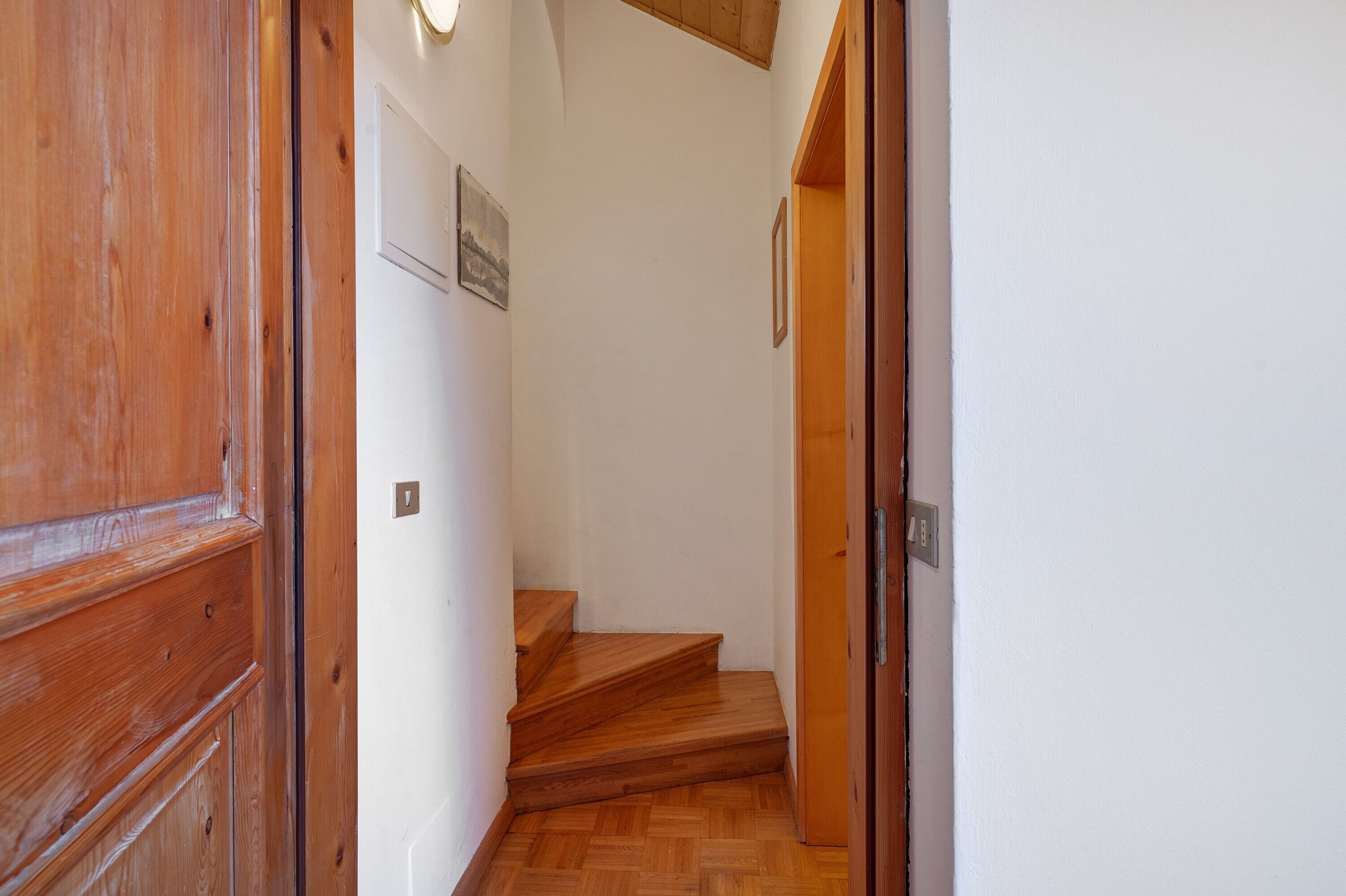 Un escalier en bois mène à une pièce aux murs blancs, où est accroché un tableau encadré.