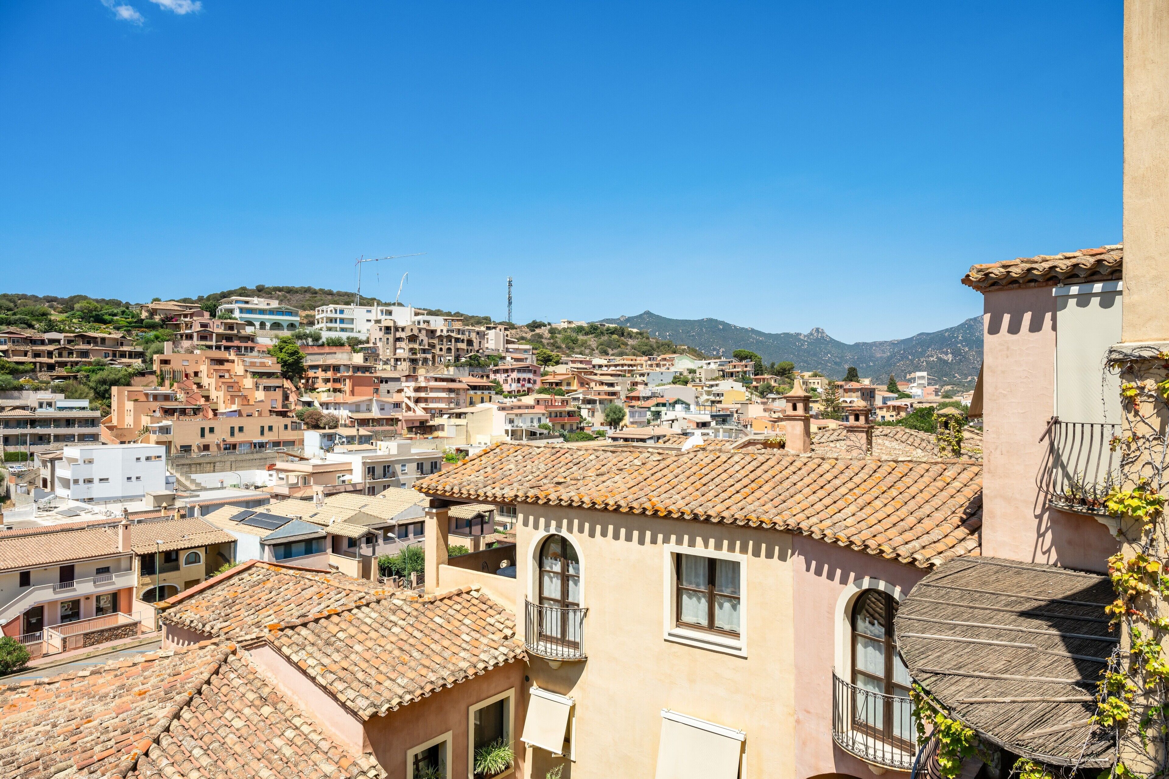 A hillside residential area with terracotta-tiled roofs, a mix of building styles, and a clear blue sky.