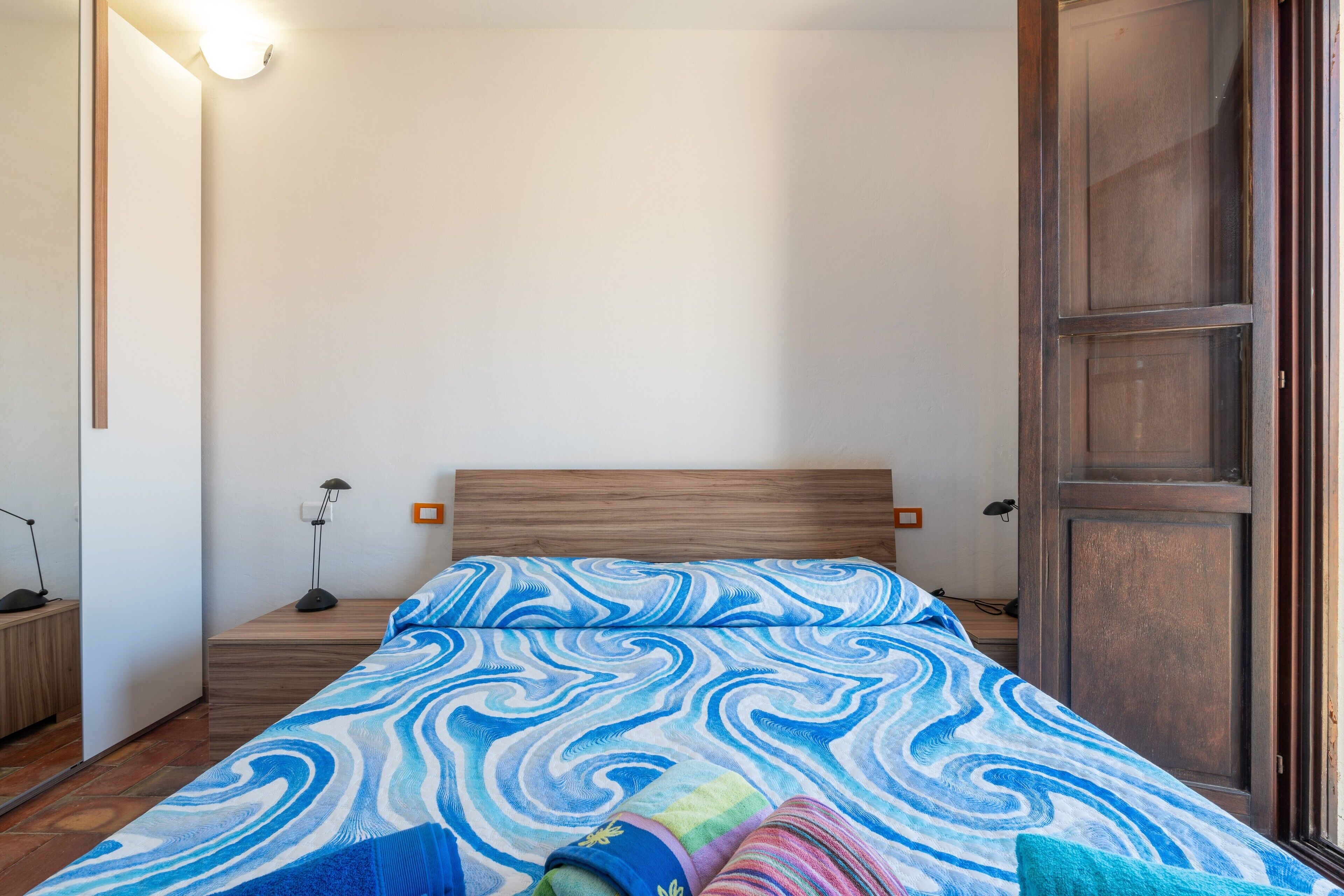 Une chambre à coucher avec une tête de lit en bois, un lit recouvert d’une couvre-lit à motifs bleus et blancs, une table de chevet avec une lampe et une petite table de nuit avec un téléphone.