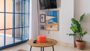 Ein Zimmer mit an der Wand montiertem Fernseher, einem runden Holztisch mit Fernbedienung, einer Topfpflanze und einem Kunstwerk an der Wand.