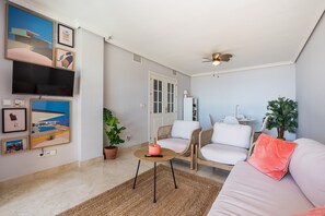 Sala de estar con sofá, sillas, una mesa de centro, televisor, obras de arte en la pared, ventilador de techo y una puerta.