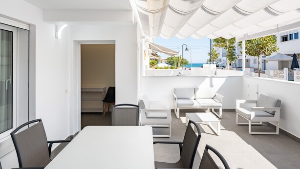Un área de estar moderna al aire libre con mobiliario blanco, una mesa y vistas al mar.