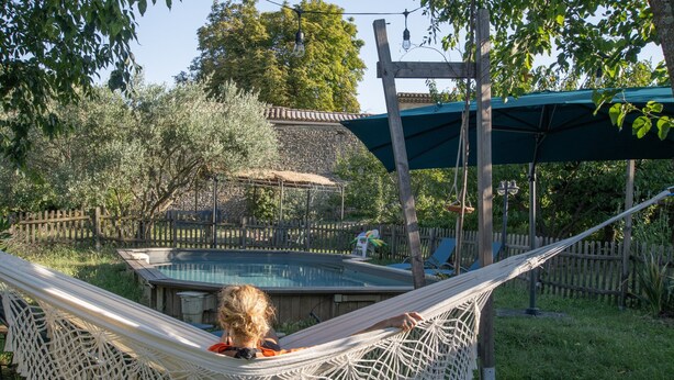 Una hamaca junto a una piscina con una terraza de madera y un edificio de piedra al fondo.