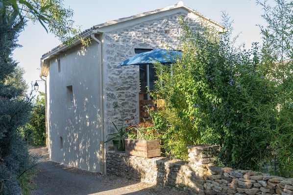 A stone building with a wooden door, a window with a blue awning, and a garden with various plants.
