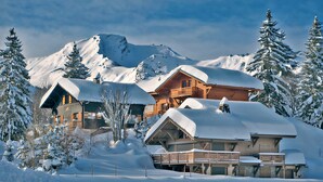 Een besneeuwde lodge met meerdere gebouwen, waaronder een hoofdgebouw en een kleinere hut, omgeven door sneeuwzware bomen en bergen.