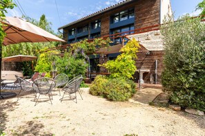 Un bâtiment à la façade en briques, doté d’un espace extérieur avec des chaises en métal et d’une pergola recouverte de plantes grimpantes.