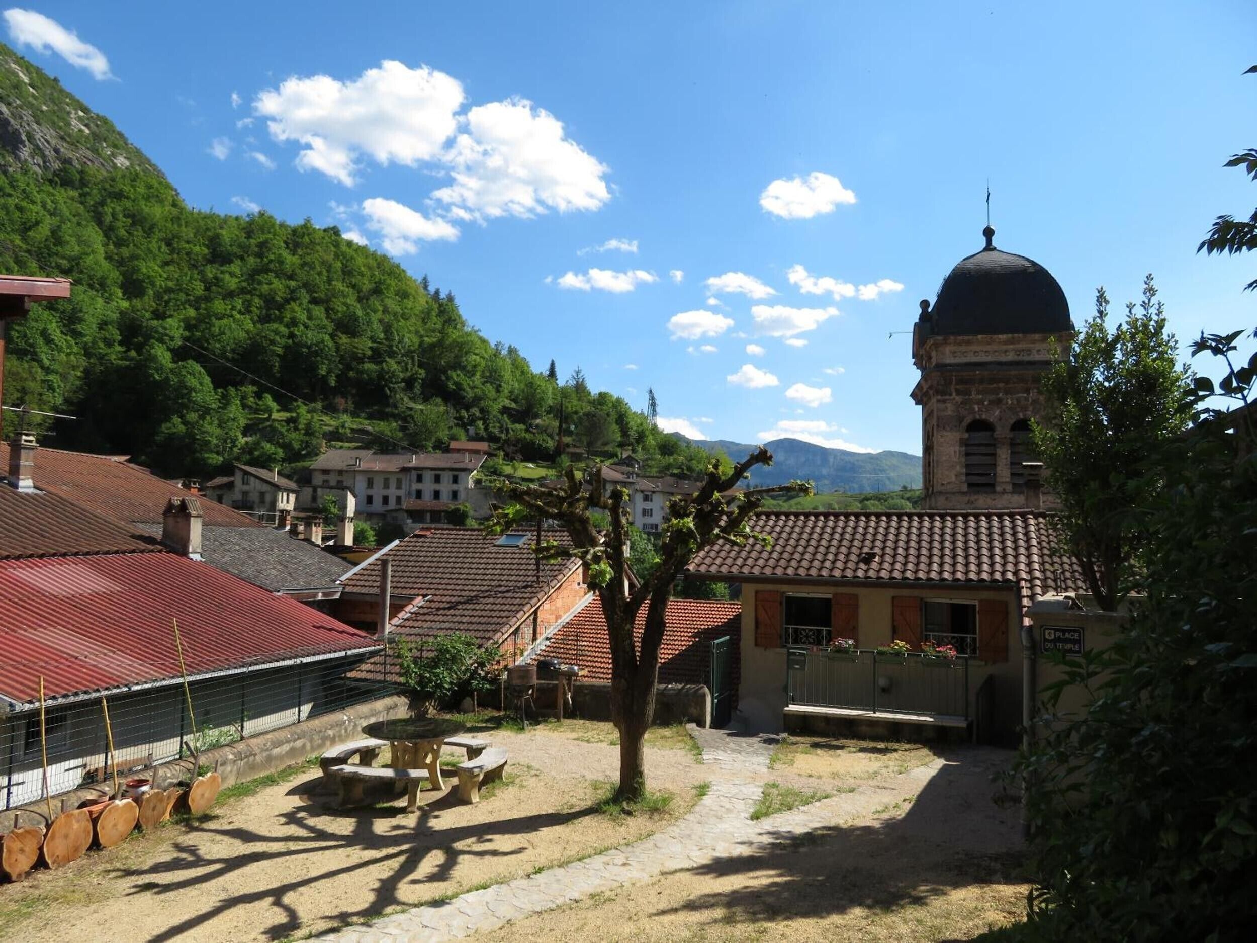 A courtyard with a stone table and benches, surrounded by buildings with red-tiled roofs and a church with a dome.