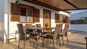 Un patio al aire libre con una mesa de madera, sillas de mimbre, una planta en maceta y una puerta con ventana.