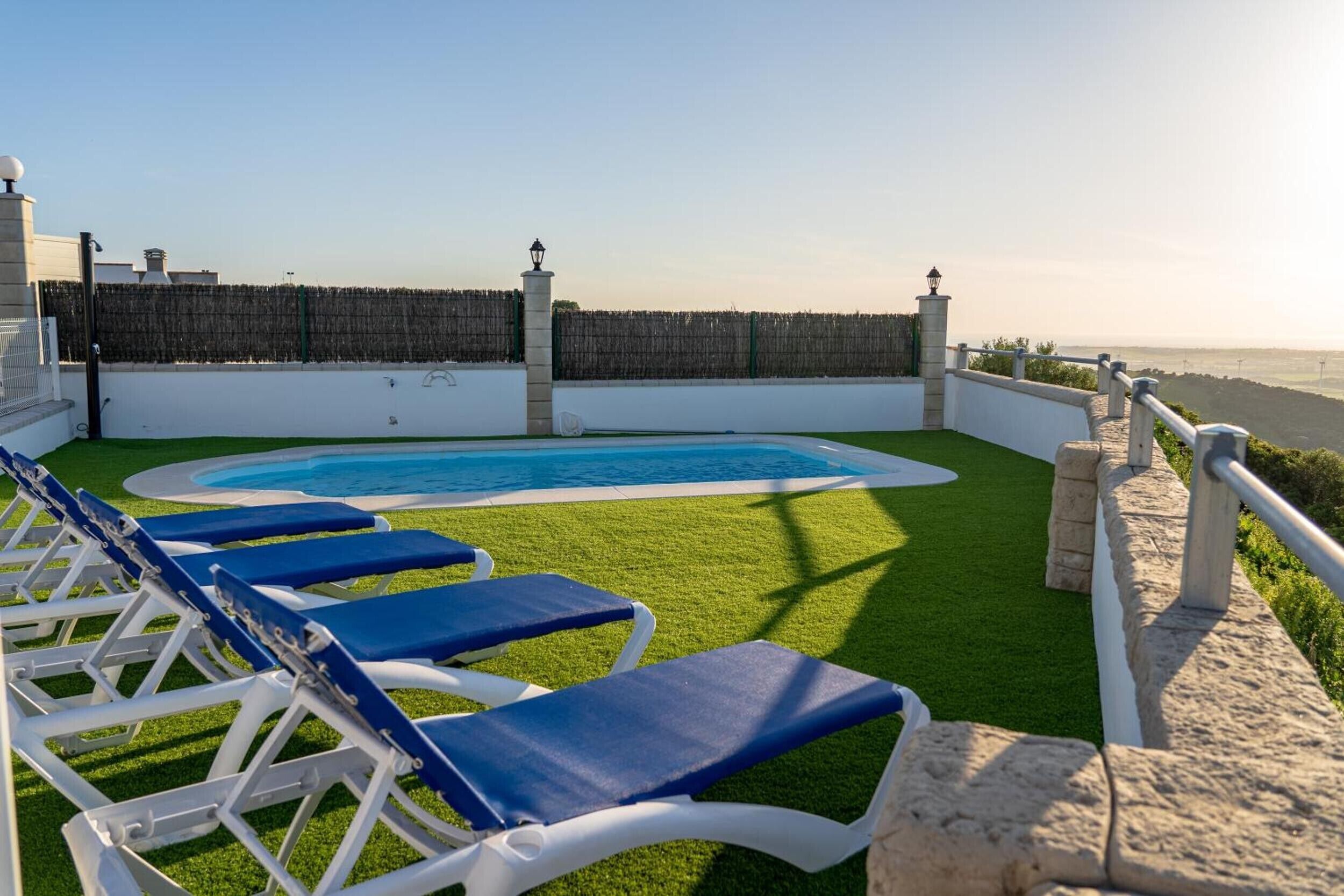 A pool area with lounge chairs, a stone wall, and a view of the sea.