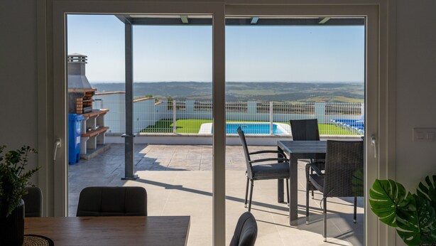 Une porte-fenêtre coulissante donne sur une terrasse extérieure équipée d’une piscine, de chaises et d’un barbecue.