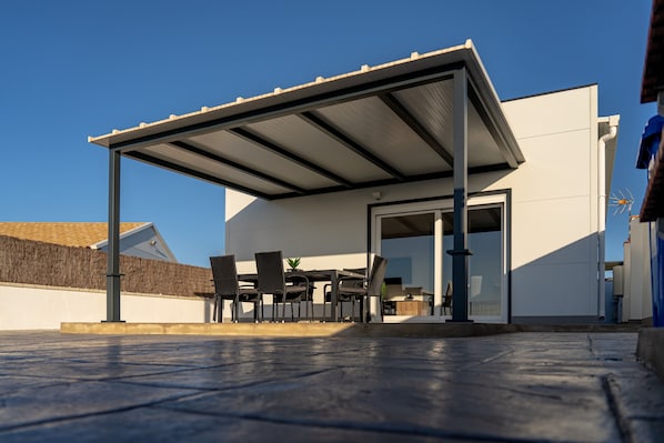 A covered outdoor seating area with a table and chairs, a potted plant, and a modern building in the background.