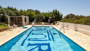 Une piscine rectangulaire aux motifs géométriques bleus, entourée de carreaux de pierre, de chaises longues blanches et d’une structure en forme de pergola.