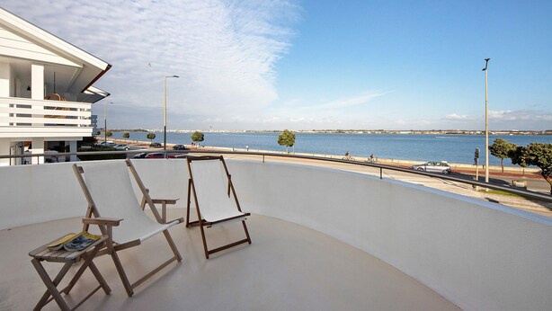 Un balcone con due poltrone in legno, vista sul mare e un edificio moderno sullo sfondo.