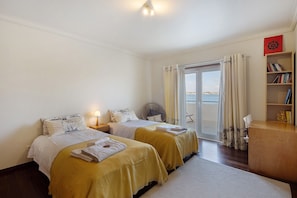 A hotel room with two beds, a wooden desk, a bookshelf, and a view of the sea through a sliding door. - Villa 'Casa Das Dunas' with Sea View, Private Terrace and Wi-Fi (Gafanha da Encarnação)