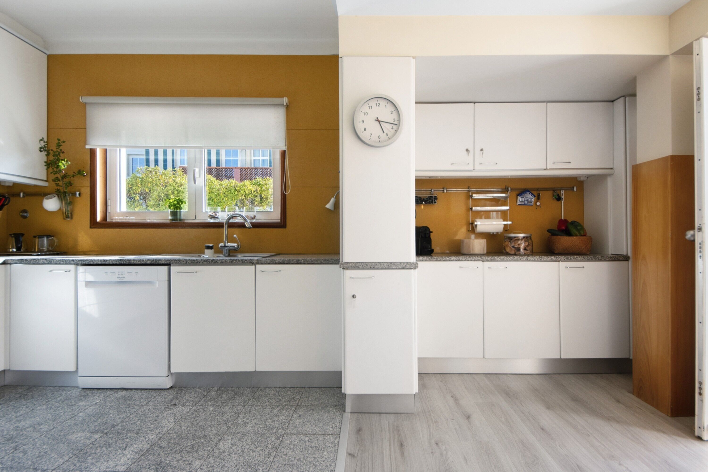 Una cucina moderna con mobili bianchi, lavastoviglie, orologio e finestra vista verde.