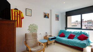 Una habitación con cama, una silla, una mesita, una planta y ventana con vista a la ciudad.