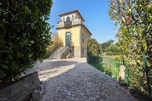 A stone building with a staircase leading to a door, surrounded by greenery and a cobblestone path.