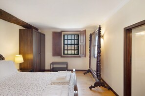 A bedroom with a bed, a nightstand, a lamp, a window with shutters, and a mirror.