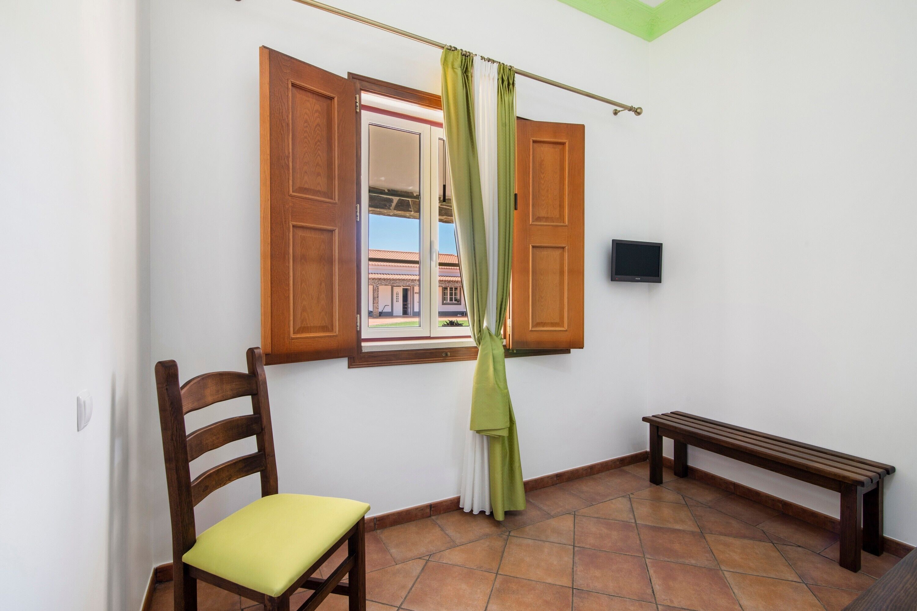 A room with a wooden chair, a bench, a window with open shutters, and a television on the wall.