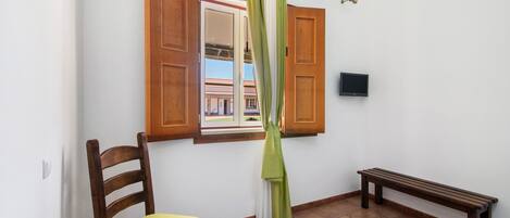 A room with a wooden chair, a bench, a window with open shutters, and a television on the wall.