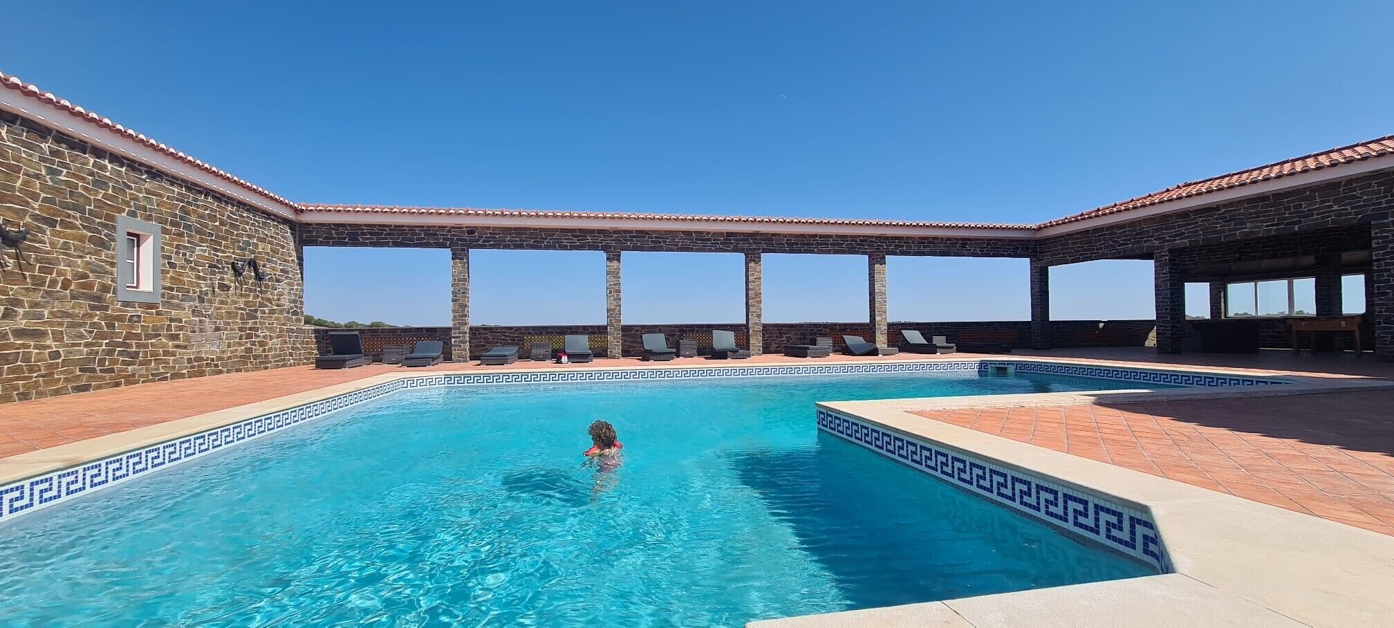 A swimming pool with stone walls and a tiled deck, surrounded by lounge chairs and a pergola.