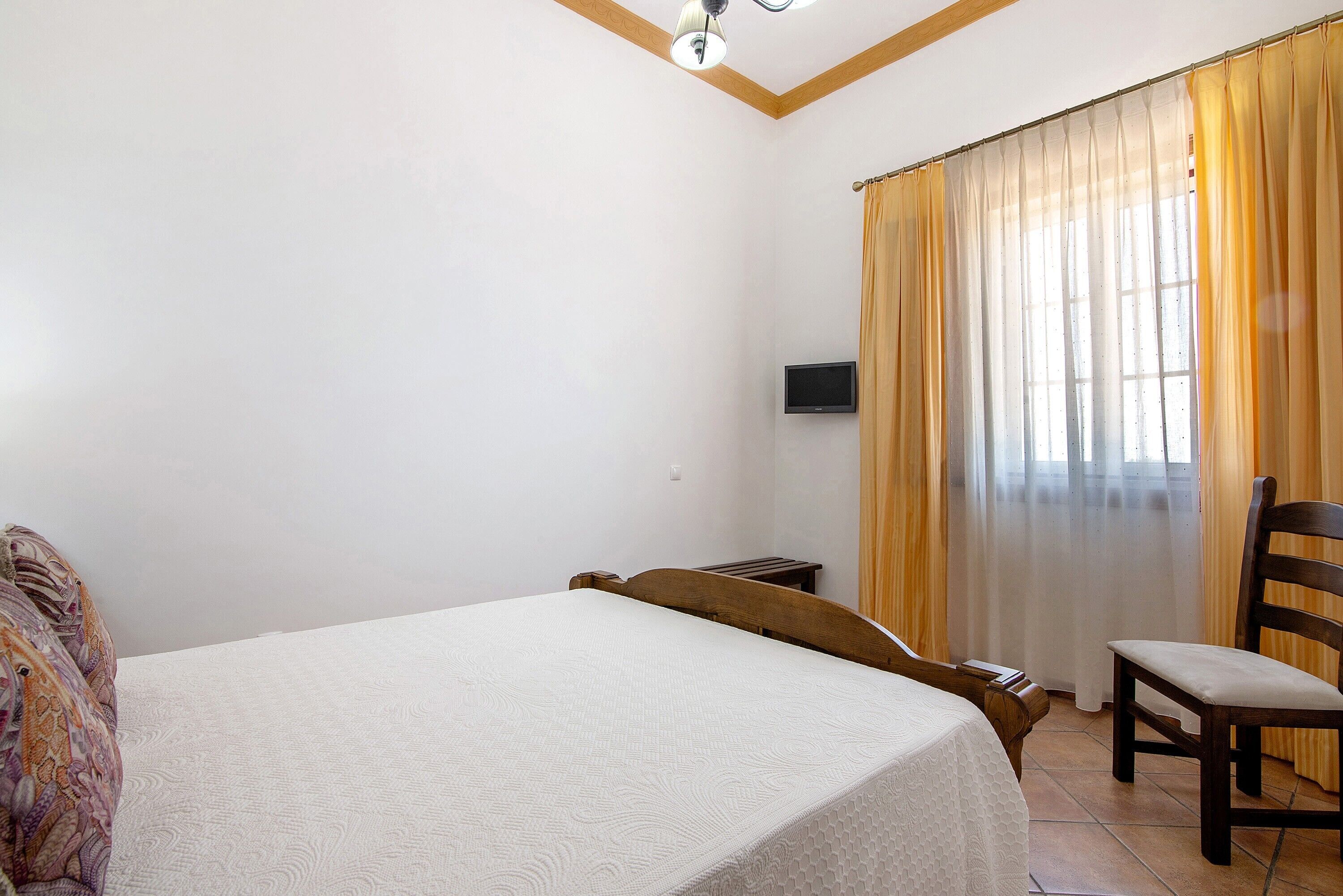 Un dormitorio con cama, una silla, un televisor en la pared y una ventana con cortinas.