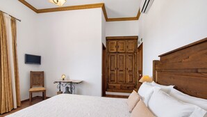 A bedroom with a large bed, a wooden wardrobe, a television, a chair, and a small table with a lamp.