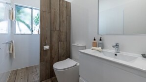 Ein modernes Badezimmer mit einer Wandpaneele aus Holz, einer weißen Toilette, einem Waschbecken mit Spiegel und einem Fenster mit Blick auf Palmen.