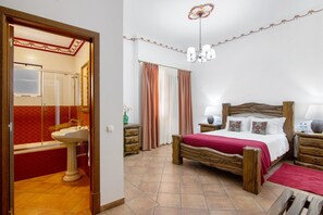 Un dormitorio con cama de madera, mesita de noche con lámpara, espejo, y baño con lavamanos y ducha.