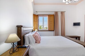 A hotel room with a bed, a bedside table with a lamp, a television, and a window with curtains. - Room 'Quarto Rosa - Countryside View' with Shared Pool, Wi-Fi and Air Conditioning (Moinhos de vento de cima)