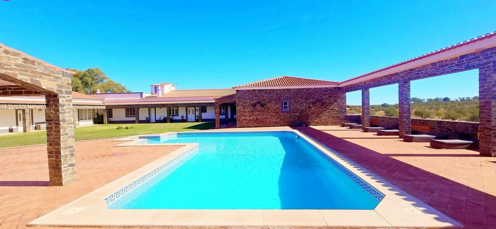 A swimming pool with a stone deck, a covered patio area, and a building with a tiled roof.