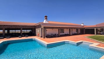 A swimming pool with a geometric tile border, a covered patio area with seating, and a clear view of the surrounding landscape.