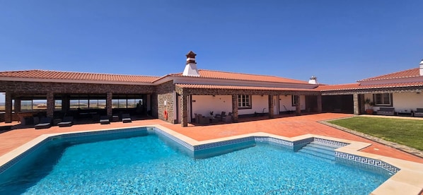 A swimming pool with a geometric tile border, a covered patio area with seating, and a clear view of the surrounding landscape.