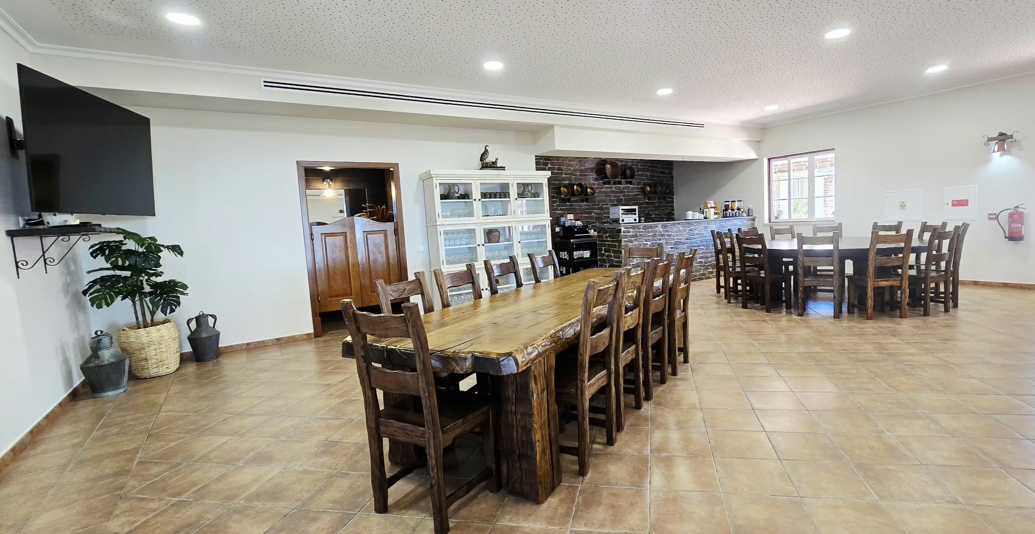 Een ruime eetruimte met een lange houten tafel en stoelen, een keuken met een achterwand van steen, en een bevestigde televisie.