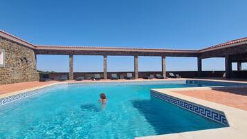 Una piscina con paredes de piedra y un deck de baldosas, rodeada de sillas de descanso y una pérgola.
