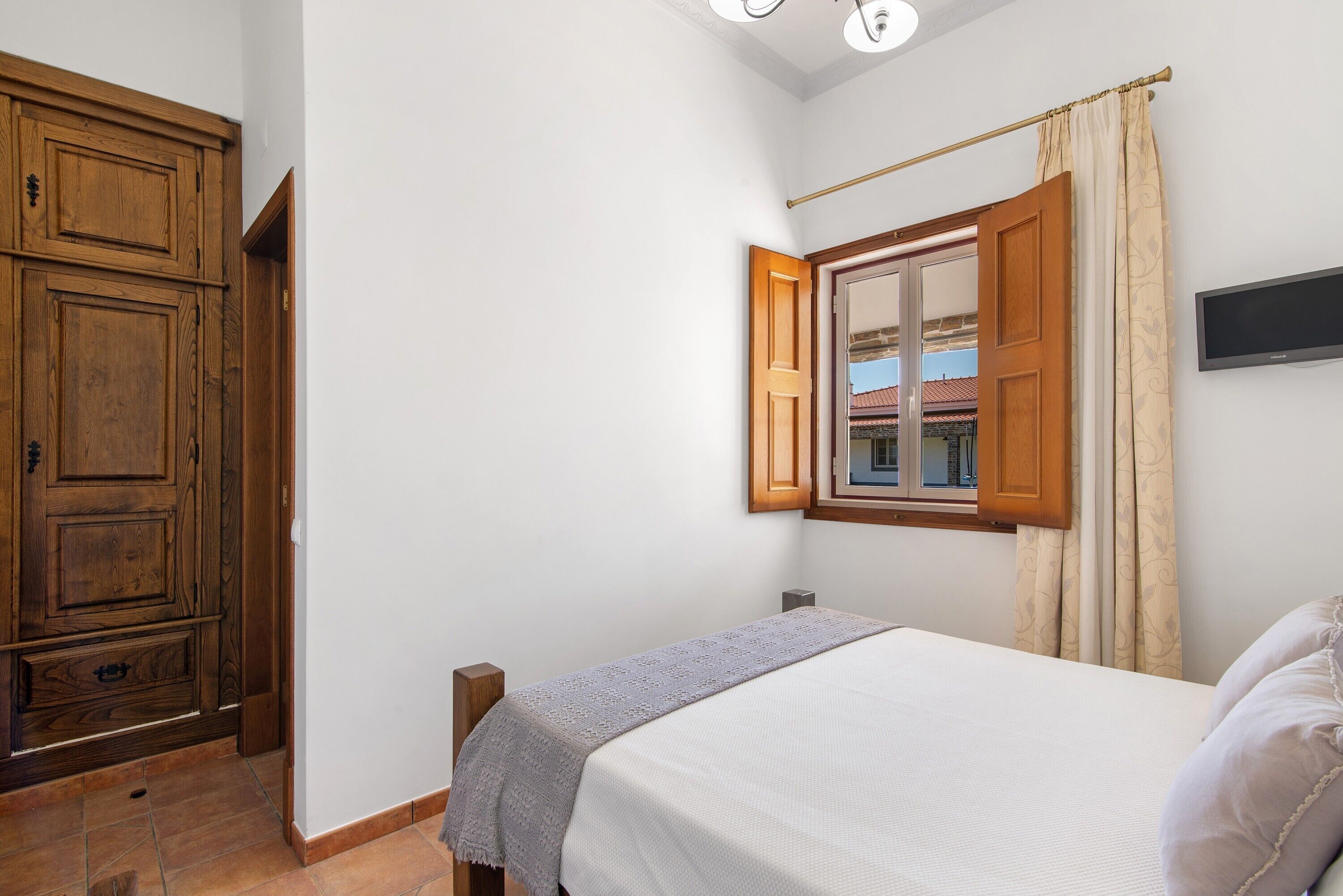 Un dormitorio con una cama, un armario de madera, una ventana con cortinas y un televisor en la pared.