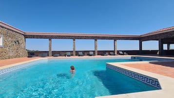 Una piscina con paredes de piedra y un deck de baldosas, rodeada de sillas de descanso y una pérgola.
