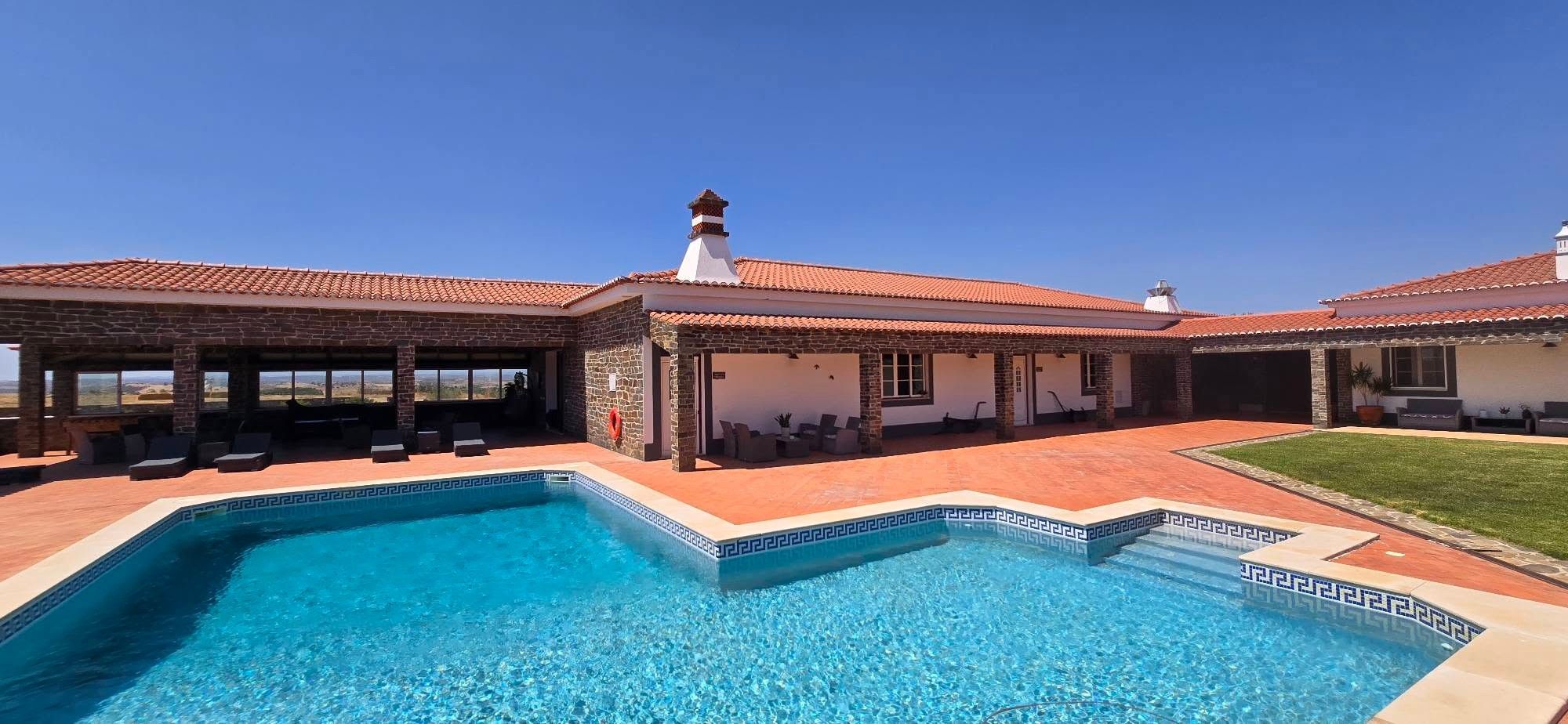 A swimming pool with a geometric tile border, a covered patio area with seating, and a clear view of the surrounding landscape.
