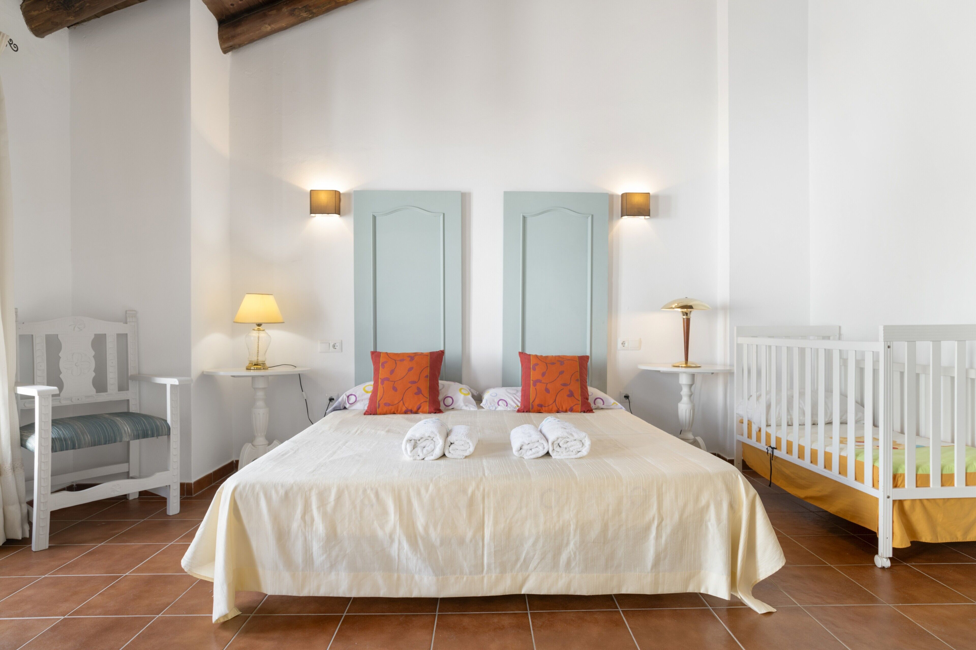A bedroom with a large bed, two bedside tables with lamps, a small chair, and a crib.
