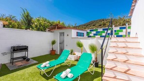 Un patio con dos sillones de exterior verdes, una parrilla de barbacoa y una escalera que conduce a un edificio con un muro de azulejos de colores.